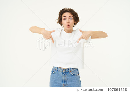 Portrait of amazed girl, pointing fingers down, showing something and looking surprised, standing over white studio background 107861838