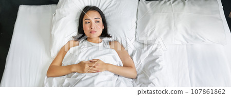 Portrait of asian girl lying alone in bed with white sheets bedding, looking thoughtful. Woman in her bedroom thinking of smth and gazing at window 107861862