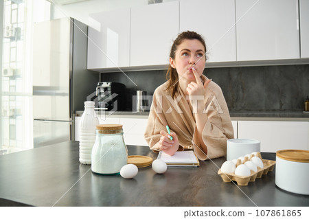 Attractive young cheerful girl baking at the kitchen, making dough, holding recipe book, having ideas Attractive young cheerful girl baking at the kitchen, making dough, holding recipe book, having ideas 107861865