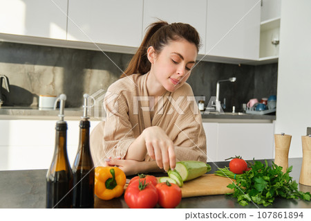 Portrait of cute young woman in bathrobe, cooking meal, standing near chopping board with vegetables, holding zucchini, making salad or vegetarian dinner 107861894