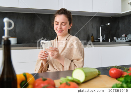 Woman watching recipe on smartphone, sitting in the kitchen with vegetables and chopping board, preparing dinner, healthy salad, cooking at home, wearing bathrobe Woman watching recipe on smartphone, sitting in the kitchen with vegetables and chopping board, preparing dinner, healthy salad, cooking at home, wearing bathrobe 107861946