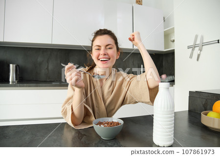 Portrait of enthusiastic young woman eating cereals with milk, looking excited and happy, sitting near kitchen worktop and having breakfast, raising hand up in triumph Portrait of enthusiastic young woman eating cereals with milk, looking excited and happy, sitting near kitchen worktop and having breakfast, raising hand up in triumph 107861973