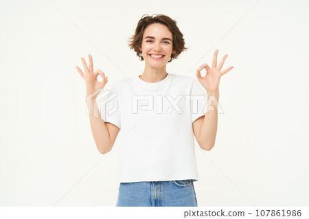 Portrait of young woman, showing okay, ok gestures and smiling, giving positive feedback, recommending, standing over white background 107861986