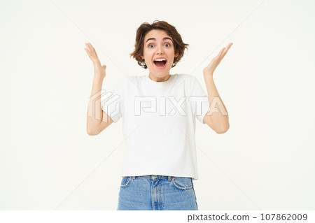 Portrait of amazed, happy young woman, raising hands up and looks surprised, standing excited against white background 107862009