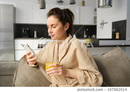 Lifestyle and morning concept. Beautiful woman sitting on sofa and resting, reading news on smartphone, drinking orange juice at home Lifestyle and morning concept. Beautiful woman sitting on sofa and resting, reading news on smartphone, drinking orange juice at home 107862014
