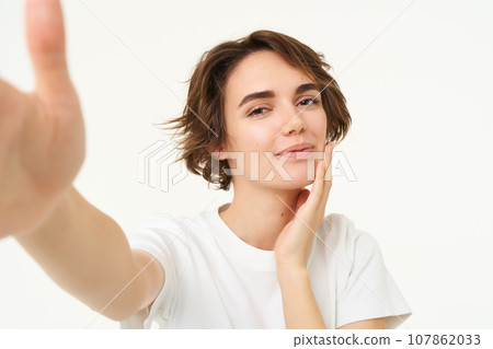 Lifestyle and people. Young woman smiling and taking selfie, posing for photo, holding camera with one hand, standing over white background 107862033