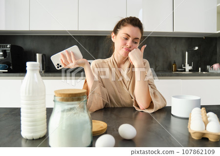 Attractive young cheerful girl baking at the kitchen, making dough, holding recipe book, having ideas Attractive young cheerful girl baking at the kitchen, making dough, holding recipe book, having ideas 107862109