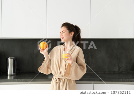 Portrait of attractive young woman, posing with an orange, drinking fresh juice at home in kitchen, starting her morning with healthy drink 107862111