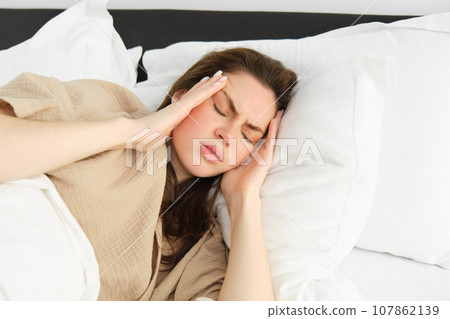 Woman lying in bed and feeling unwell, having headache, touching her head with frustrated face, has high fever or migraine. concept of health and people 107862139