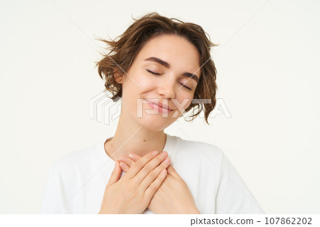 Close up portrait of young woman feeling warmth and love, holds hands on heart and smiles, stands over white studio background Close up portrait of young woman feeling warmth and love, holds hands on heart and smiles, stands over white studio background 107862202