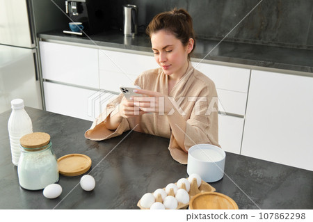 Attractive young cheerful girl baking at the kitchen, making dough, holding recipe book, having ideas 107862298