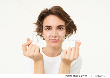 Close up of brunette woman shows finger hearts gesture and smiles, isolated over white background 107862328