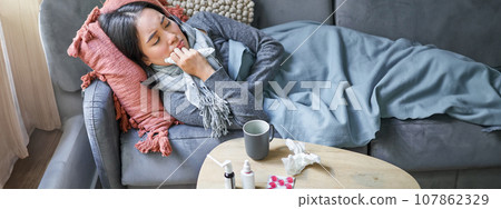 Sick sad korean woman lying on sofa, feeling unwell, catching cold, flu and temperature, looking upset, taking medication 107862329