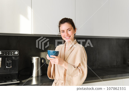 Daily routine and lifestyle. Young beautiful woman in bathrobe, standing in kitchen with cup of coffee, drinking tea, smiling and looking happy 107862331