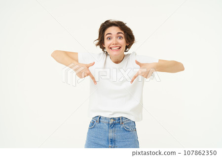 Portrait of cute, happy young woman pointing fingers down, showing advertisement, demonstrating banner on the bottom, standing against white background 107862350