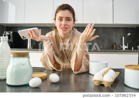 Attractive young cheerful girl baking at the kitchen, making dough, holding recipe book, having ideas Attractive young cheerful girl baking at the kitchen, making dough, holding recipe book, having ideas 107862354