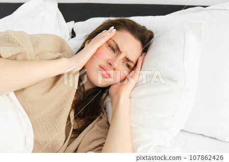 Close up portrait of sick woman lying in her bed and feeling unwell, touching head, has headache, looking frustrated from migraine, wearing pyjamas 107862426