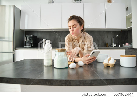 Attractive young cheerful girl baking at the kitchen, making dough, holding recipe book, having ideas Attractive young cheerful girl baking at the kitchen, making dough, holding recipe book, having ideas 107862444