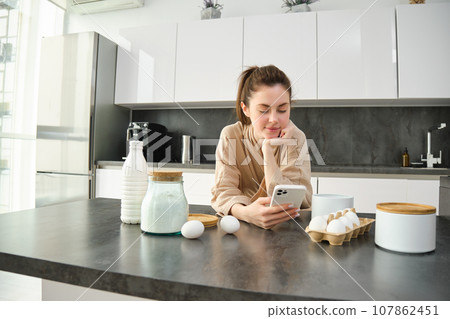 Attractive young cheerful girl baking at the kitchen, making dough, holding recipe book, having ideas 107862451