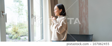 Beautiful korean woman staying at home, looking outside window, drinking coffee espresso and smiling, feeling comfort and warmth Beautiful korean woman staying at home, looking outside window, drinking coffee espresso and smiling, feeling comfort and warmth 107862455