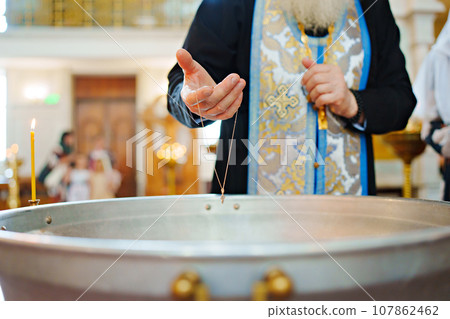 The priest's hand dips a chain with a cross into the water of the baptismal font 107862462
