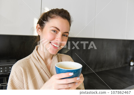 Close up portrait of beautiful, smiling woman, holding cup of coffee, posing in the kitchen, enjoying her morning at home, wearing bathrobe Close up portrait of beautiful, smiling woman, holding cup of coffee, posing in the kitchen, enjoying her morning at home, wearing bathrobe 107862467