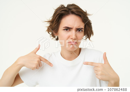 Close up portrait of woman with confused face, looks offended and points at herself, stands over white studio background Close up portrait of woman with confused face, looks offended and points at herself, stands over white studio background 107862488
