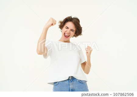 Cheerful brunette woman winning, triumphing, celebrating victory, achieves goal, stands over white background 107862506