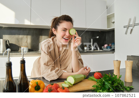 Portrait of brunette woman, wife cooking at home, making dinner, posing near chopping board in kitchen with vegetables, holding zucchini Portrait of brunette woman, wife cooking at home, making dinner, posing near chopping board in kitchen with vegetables, holding zucchini 107862521