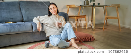 Portrait of stylish korean woman sits on floor with smartphone, using mobile phone, smiling pleased, concept of staying at home and relaxation Portrait of stylish korean woman sits on floor with smartphone, using mobile phone, smiling pleased, concept of staying at home and relaxation 107862616