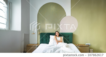 Vertical shot of beautiful asian girl lying in bedroom with sophisticated interrior, smiling and looking happy in morning, spending time in bed Vertical shot of beautiful asian girl lying in bedroom with sophisticated interrior, smiling and looking happy in morning, spending time in bed 107862617