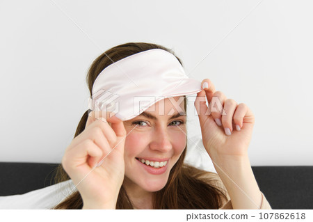 Close up portrait of gorgeous smiling woman with sleeping mask on forehead, waking up in morning, relaxing in bed, laughing and looking happy, resting on vacation in hotel room Close up portrait of gorgeous smiling woman with sleeping mask on forehead, waking up in morning, relaxing in bed, laughing and looking happy, resting on vacation in hotel room 107862618