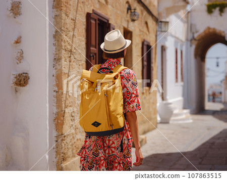 Nice asian Female in boho dress Enjoying Sunny Day on Greek Islands. Travel to Lindos, Mediterranean islands tourist season. Tourist woman on vacation in Greece walking through the streets of Lindos. 107863515