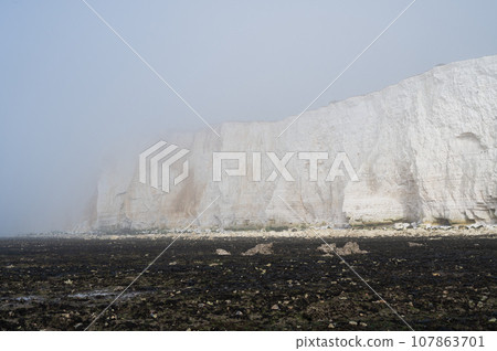 Misty morning in Hope Gap beach, Cuckmere Haven, located between Seaford and Eastbourne. Pebbly coastline with seaweed and the white cliffs on the background, selective focus 107863701