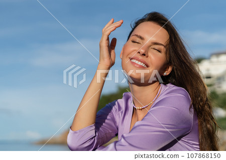 A young beautiful woman in a romantic mood, lilac silk clothes, on the beach against the backdrop of the sea and stones at sunset, laughs, smiles with a snow-white smile 107868150