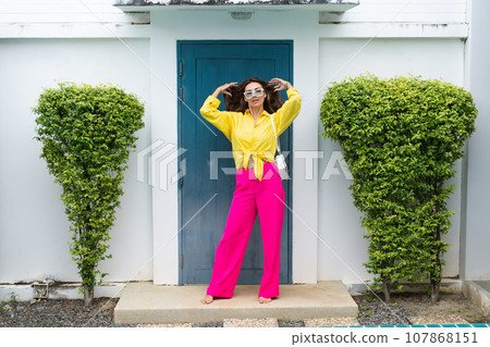 Stylish fit fashion women in bright pink wide leg pants and yellow shirt holding bag trendy mint sunglasses posing at luxury tropical villa by pool outdoor natural day light Stylish fit fashion women in bright pink wide leg pants and yellow shirt holding bag trendy mint sunglasses posing at luxury tropical villa by pool outdoor natural day light 107868151
