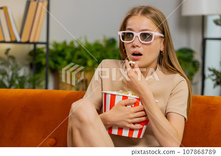 Woman sitting on couch eating popcorn and watching interesting TV 3D film, sport game online at home 107868789
