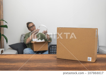 Attentive female consumer inspects new headphones near table with box of goods in light housing inspection of arrived goods and cardboard packaging with barcode sticker 107868817