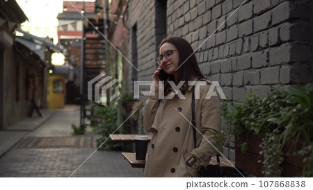 A young woman talks on the phone while standing against a wall on a narrow street. A girl with glasses and a coat speaks on the phone. 107868838
