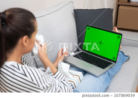 Young woman wipes nose with handkerchief talking to therapist on online call on laptop lying on sofa patient with influenza receives support of doctor remotely from home side view Young woman wipes nose with handkerchief talking to therapist on online call on laptop lying on sofa patient with influenza receives support of doctor remotely from home side view 107869039