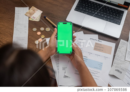 Mobile Financial Control: A woman's hand holds a mobile phone with a chroma key screen on a wooden desk, accompanied by utility bills, cash, and an online banking app.  107869386
