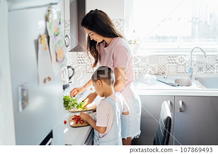 Loving young mother teaching her preschooler son prepare healthy food or salad at home kitchen, caring happy mom cooking together with kid on modern kitchen, boy involved in preparing food with mum Loving young mother teaching her preschooler son prepare healthy food or salad at home kitchen, caring happy mom cooking together with kid on modern kitchen, boy involved in preparing food with mum 107869493