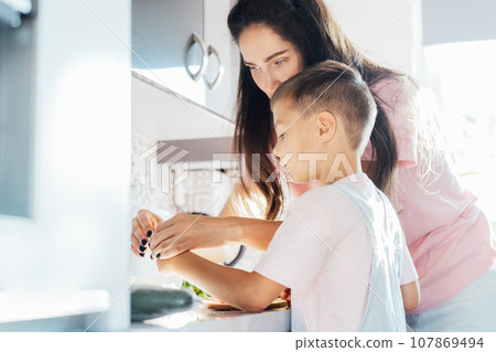 Caring young Mother putting a plaster on child finger with wound on from a cut by kitchen knife. First aid in case of small domestic injury. Negligence in cooking at home. Caring young Mother putting a plaster on child finger with wound on from a cut by kitchen knife. First aid in case of small domestic injury. Negligence in cooking at home. 107869494