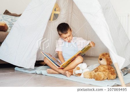 Cute little preschooler boy reading book to his toys sitting in cozy kids teepee tent in the living room at home. Child entertaining himself. Home children's activity 107869495