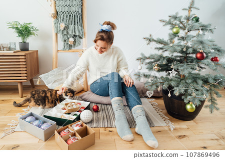 Young woman in cozy sweater petting her cat while decorating potted Christmas tree in light modern Scandinavian interior home. Eco-friendly winter holidays. Domestic pet at home. Selective focus. Young woman in cozy sweater petting her cat while decorating potted Christmas tree in light modern Scandinavian interior home. Eco-friendly winter holidays. Domestic pet at home. Selective focus. 107869496