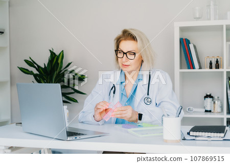 Friendly female middle aged doctor shows jar of pills and pill organizer to laptop in clinic office. Video call online, treatment remote, pharmacy, supplement advice, medical healthcare. Telemedicine 107869515