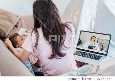 Back view mother measures temperature of the child with non-contact digital infrared thermometer during online video chat with pediatrician doctor on pc. Telemedicine, telehealth. Cold season concept. 107869552