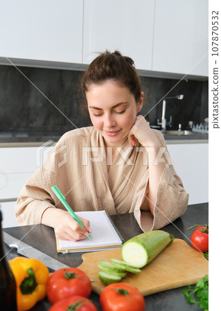 Vertical shot of smiling woman making notes, writing cooking recipe, creates groceries list, sits in the kitchen with vegetables and chopping board 107870532
