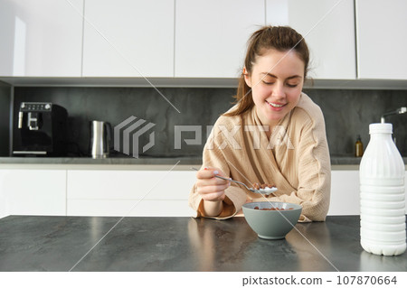 Portrait of happy young woman leans on kitchen worktop and eating cereals, has milk and bowl in front of her, having her breakfast, wearing bathrobe Portrait of happy young woman leans on kitchen worktop and eating cereals, has milk and bowl in front of her, having her breakfast, wearing bathrobe 107870664