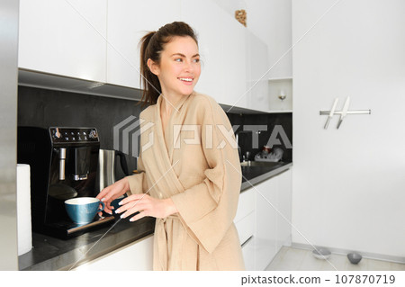 Portrait of modern woman in the morning, standing in the kitchen and making herself cup of coffee, turn around and smiling, wearing bathrobe, starting her day with delicious cappuccino Portrait of modern woman in the morning, standing in the kitchen and making herself cup of coffee, turn around and smiling, wearing bathrobe, starting her day with delicious cappuccino 107870719
